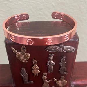 May include: A copper-coloured cuff bracelet with engraved circular details. The open-ended bracelet is displayed on a dark wooden stand adorned with small, decorative metal figures. The bracelet's design is simple and elegant.
