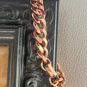 May include: A rose gold chain bracelet with large, interlocking links. The bracelet is displayed against a dark, ornate picture frame. The chain has a polished finish and a secure clasp.