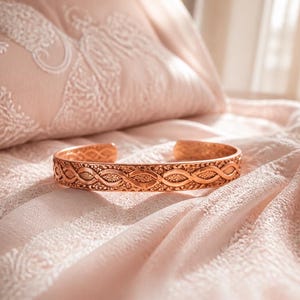 Peut inclure: Un bracelet manchette en cuivre présentant un motif gaufré complexe de boucles tressées répétitives et de détails texturés. La bande métallique possède un design ouvert, mettant en valeur une finition brun-rouge métallique et un travail soigné.