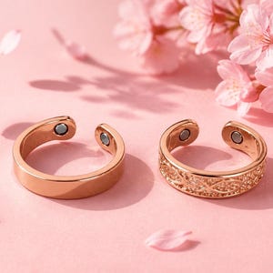 May include: Two rose gold-toned open rings with magnetic inserts at the ends. One ring features a smooth, plain surface, while the other displays an intricate, repeating infinity-style pattern across the outer band. Both rings are circular.