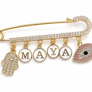 May include: A gold-tone safety pin brooch featuring a row of clear crystals along the top bar. Dangling from the pin are four circular white charms spelling MAYA in gold letters, a crystal-encrusted hamsa hand charm, and a pink crystal evil eye charm.