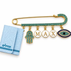 May include: A gold-tone safety pin brooch adorned with turquoise crystals, featuring dangling charms: a hamsa hand, three white circular discs with the letters M, A, and X, and an evil eye charm. A small, light blue book with Hebrew text is attached.