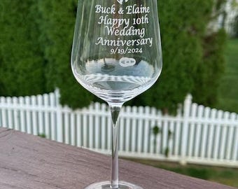 May include: A clear wine glass with a long stem and a round base. The glass is engraved with the words "Buck & Elaine Happy 10th Wedding Anniversary 9/19/2024" and features a bell and ring design. The glass is sitting on a wooden surface.