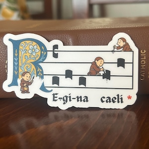 Puede incluir: Una pegatina con un pentagrama musical y monjes de dibujos animados. El diseño incluye una letra "R" azul adornada con flores y la frase latina "Ergi-na caeli". Los monjes visten túnicas marrones.