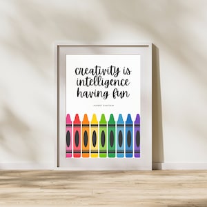 Puede incluir: Una impresión enmarcada en blanco con la cita "La creatividad es inteligencia teniendo diversión" de Albert Einstein. La cita está en texto negro y está rodeada de una fila de crayones de colores.