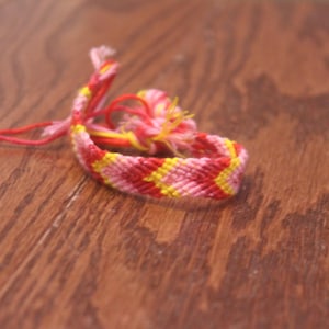 May include: A pink and yellow friendship bracelet with a woven pattern. The bracelet is made of string and is tied in a knot.