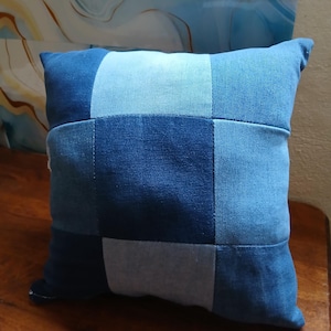 Pode incluir: Almofada quadrada de ganga com um design de patchwork em vários tons de azul. A almofada apresenta uma forma retangular com um detalhe de costura. A almofada é feita de tecido de ganga.