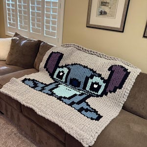 Puede incluir: Una manta de punto grueso con un dise&ntilde;o pixelado del personaje Stitch de Lilo & Stitch. La manta es principalmente gris claro con detalles en azul, morado y negro. Est&aacute; sobre un sof&aacute; marr&oacute;n.