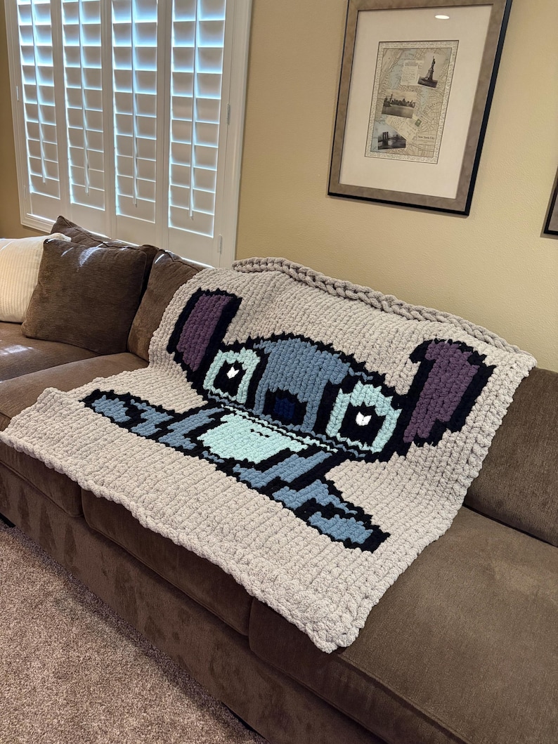 Puede incluir: Una manta de punto grueso con un dise&ntilde;o pixelado del personaje Stitch de Lilo & Stitch. La manta es principalmente gris claro con detalles en azul, morado y negro. Est&aacute; sobre un sof&aacute; marr&oacute;n.