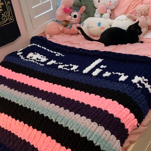 K&ouml;nnte beinhalten: Eine grob gestrickte Decke mit abwechselnden Streifen in Marineblau, Rosa, Schwarz, Lila und Hellblau. Die Decke tr&auml;gt den Namen "Olivia" in wei&szlig;er Wolle. Die Decke liegt auf einem Bett mit Stofftieren.