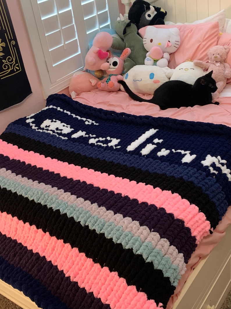 K&ouml;nnte beinhalten: Eine grob gestrickte Decke mit abwechselnden Streifen in Marineblau, Rosa, Schwarz, Lila und Hellblau. Die Decke tr&auml;gt den Namen "Olivia" in wei&szlig;er Wolle. Die Decke liegt auf einem Bett mit Stofftieren.