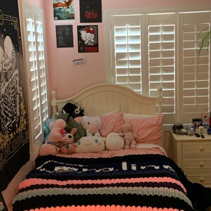 K&ouml;nnte beinhalten: Ein Schlafzimmer mit wei&szlig;em Bettrahmen, rosa W&auml;nden und wei&szlig;en Fensterl&auml;den. Das Bett ist mit einer gestreiften marineblauen, rosa und grauen Decke und mehreren Pl&uuml;schtieren bedeckt. An der Wand h&auml;ngen Poster, darunter ein Hello Kitty "Psycho" Poster.