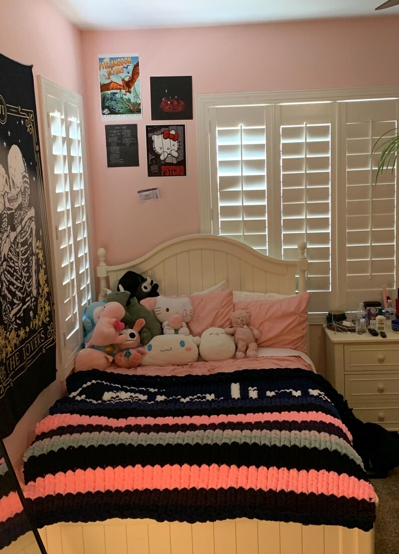 K&ouml;nnte beinhalten: Ein Schlafzimmer mit wei&szlig;em Bettrahmen, rosa W&auml;nden und wei&szlig;en Fensterl&auml;den. Das Bett ist mit einer gestreiften marineblauen, rosa und grauen Decke und mehreren Pl&uuml;schtieren bedeckt. An der Wand h&auml;ngen Poster, darunter ein Hello Kitty "Psycho" Poster.