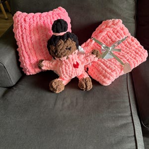 Peut inclure: Une poup&eacute;e pour b&eacute;b&eacute; rose au crochet faite &agrave; la main avec des cheveux bruns et un c&oelig;ur rouge sur la poitrine, &agrave; c&ocirc;t&eacute; d'une couverture et d'un oreiller roses. La couverture est attach&eacute;e avec un ruban argent&eacute;. La poup&eacute;e est sur un canap&eacute; gris fonc&eacute;.