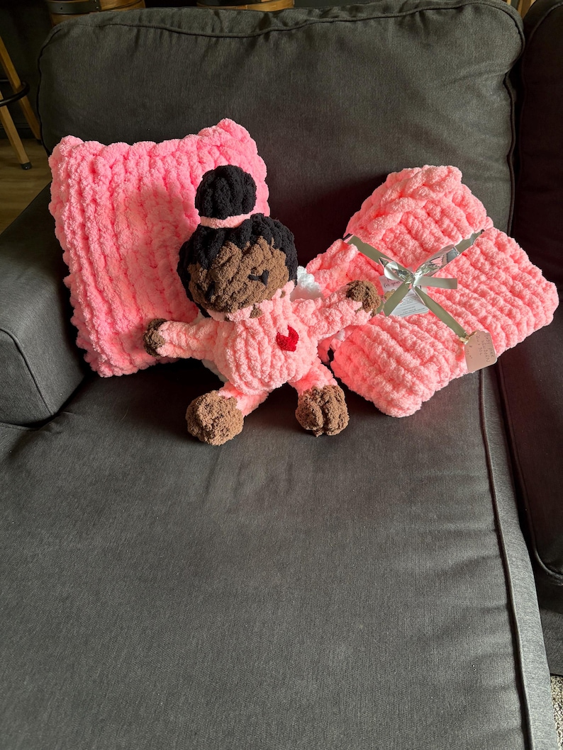 Peut inclure: Une poup&eacute;e pour b&eacute;b&eacute; rose au crochet faite &agrave; la main avec des cheveux bruns et un c&oelig;ur rouge sur la poitrine, &agrave; c&ocirc;t&eacute; d'une couverture et d'un oreiller roses. La couverture est attach&eacute;e avec un ruban argent&eacute;. La poup&eacute;e est sur un canap&eacute; gris fonc&eacute;.