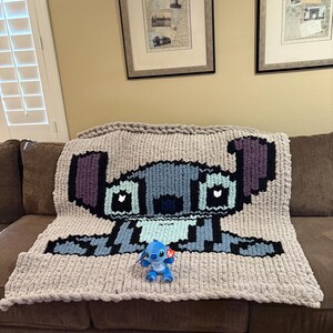 Puede incluir: Una manta de punto grueso con un dise&ntilde;o pixelado de un personaje de dibujos animados azul, colocada sobre un sof&aacute; marr&oacute;n. Un peque&ntilde;o peluche del mismo personaje est&aacute; colocado delante de la manta. La manta es principalmente gris, con detalles azules, negros y morados.