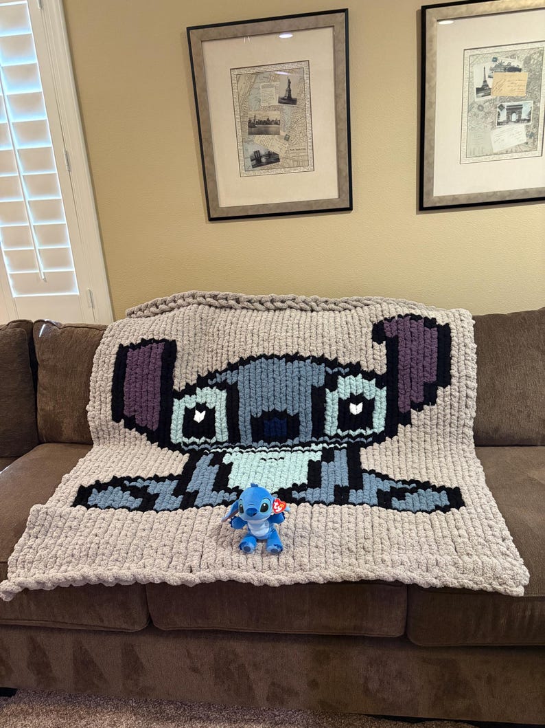 Puede incluir: Una manta de punto grueso con un dise&ntilde;o pixelado de un personaje de dibujos animados azul, colocada sobre un sof&aacute; marr&oacute;n. Un peque&ntilde;o peluche del mismo personaje est&aacute; colocado delante de la manta. La manta es principalmente gris, con detalles azules, negros y morados.