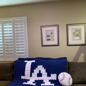 May include: A navy blue knitted blanket with a white "LA" logo, resting on a brown couch. A baseball-shaped pillow with red stitching sits beside the blanket. The blanket is a cozy home decor item.