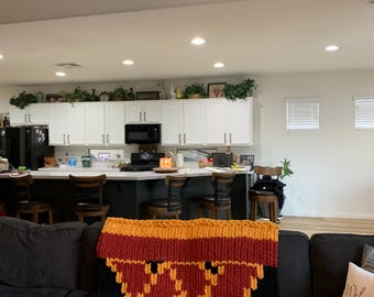 May include: A chunky knit blanket in shades of gold, red, and black, draped over a dark gray sofa. The blanket features a geometric pattern and a thick, textured weave, adding a cozy touch to the living room setting.