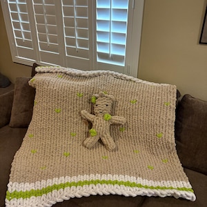 May include: A handmade, beige knitted blanket with a white and green border, and a matching stuffed animal. The blanket has green heart-shaped accents. The blanket is displayed on a brown couch.