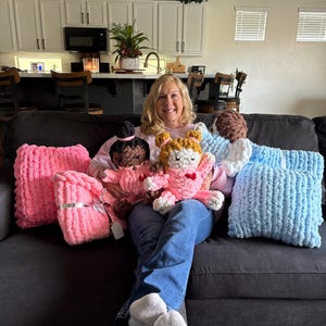 Peut inclure: Une femme assise sur un canap&eacute; gris fonc&eacute; tient plusieurs poup&eacute;es et coussins en peluche faits &agrave; la main. Les poup&eacute;es ont des cheveux bruns, noirs et oranges. Les coussins sont roses et bleus. Les poup&eacute;es et les coussins sont faits d'un fil &eacute;pais et doux.