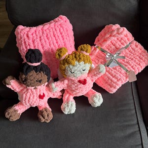 Peut inclure: Ensemble de couverture et d'oreiller pour b&eacute;b&eacute; en crochet rose, avec deux poup&eacute;es en crochet. Les poup&eacute;es ont des cheveux bruns et blonds, des visages blancs et des c&oelig;urs rouges. La couverture est attach&eacute;e avec un ruban argent&eacute;.