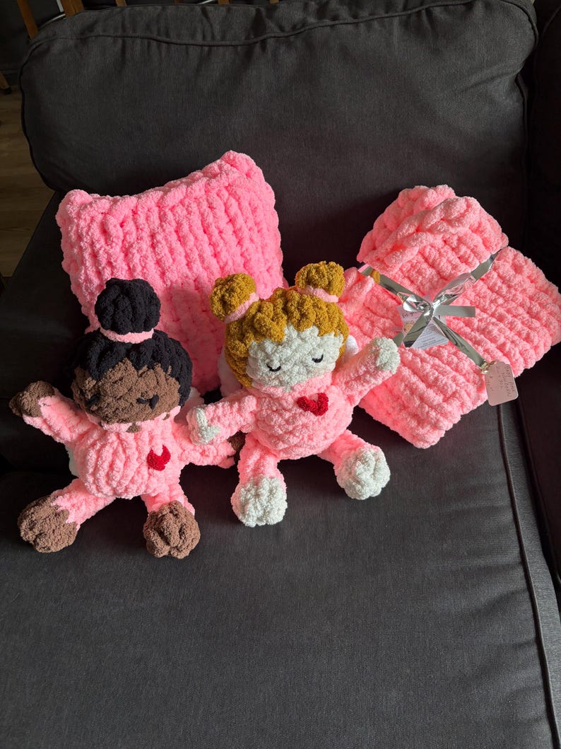 Peut inclure: Ensemble de couverture et d'oreiller pour b&eacute;b&eacute; en crochet rose, avec deux poup&eacute;es en crochet. Les poup&eacute;es ont des cheveux bruns et blonds, des visages blancs et des c&oelig;urs rouges. La couverture est attach&eacute;e avec un ruban argent&eacute;.