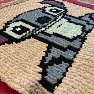 Puede incluir: Una manta grande, pixelada y hecha a mano con un dise&ntilde;o de robot en tonos grises, negros, azules y verde menta. La manta tiene un borde beige y est&aacute; hecha de un hilo grueso y suave. La manta se muestra sobre una superficie roja.
