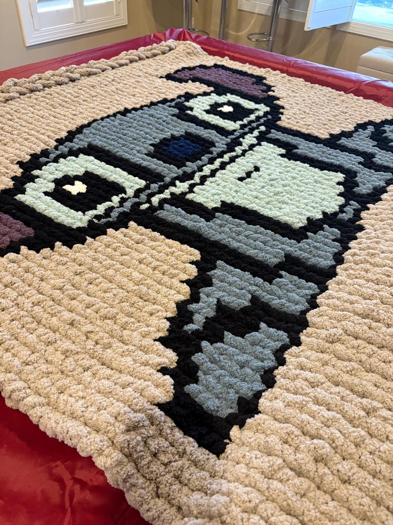 Puede incluir: Una manta grande, pixelada y hecha a mano con un dise&ntilde;o de robot en tonos grises, negros, azules y verde menta. La manta tiene un borde beige y est&aacute; hecha de un hilo grueso y suave. La manta se muestra sobre una superficie roja.