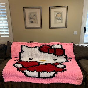 May include: A pink, chunky-knit blanket featuring a large, pixelated image of Hello Kitty. The design includes white, red, black, and gold details. The blanket is draped over a brown sofa.