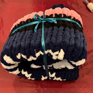 K&ouml;nnte beinhalten: Eine gefaltete, grob gestrickte Decke in Marineblau, Schwarz und Rosa, gebunden mit einem t&uuml;rkisfarbenen Band. Die Decke hat ein weiches, strukturiertes Aussehen, mit wei&szlig;en Akzenten an den R&auml;ndern. Diese gem&uuml;tliche Decke ist perfekt zum Einkuscheln.