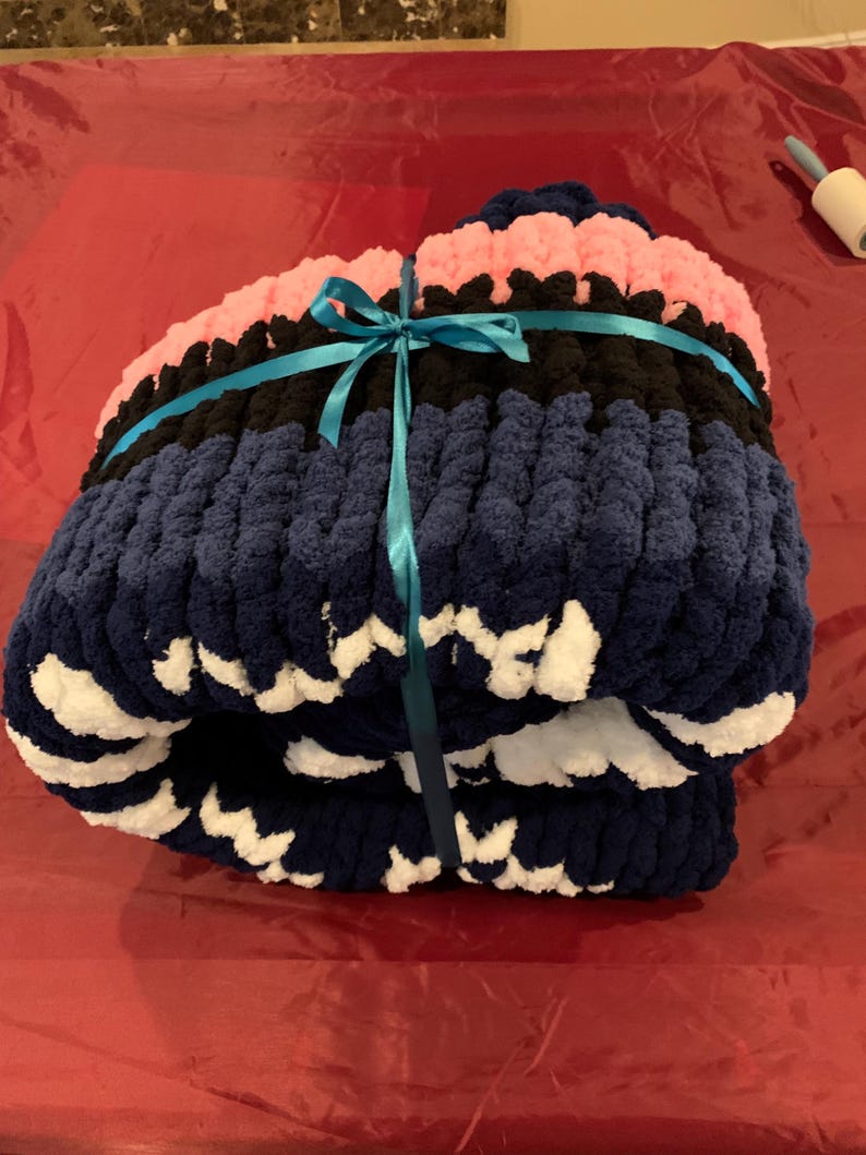K&ouml;nnte beinhalten: Eine gefaltete, grob gestrickte Decke in Marineblau, Schwarz und Rosa, gebunden mit einem t&uuml;rkisfarbenen Band. Die Decke hat ein weiches, strukturiertes Aussehen, mit wei&szlig;en Akzenten an den R&auml;ndern. Diese gem&uuml;tliche Decke ist perfekt zum Einkuscheln.