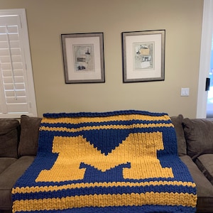 May include: A chunky knit blanket in navy blue and gold, featuring a large gold letter "M" in the center. The blanket is draped over a brown sofa. The color scheme is reminiscent of a college team.