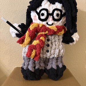 Könnte beinhalten: Eine handgefertigte Plüschpuppe mit schwarzen Haaren, runder Brille und einem rot-goldenen Schal. Die Puppe trägt einen grauen Pullover und eine graue Hose mit schwarzen Schuhen. Sie hält einen schwarzen Zauberstab mit weißer Spitze.