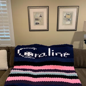 K&ouml;nnte beinhalten: Eine grob gestrickte Decke mit marineblauen, schwarzen, rosa und hellblauen Streifen. Der obere Bereich zeigt den Namen "Caroline" in wei&szlig;en Buchstaben. Die Decke liegt auf einem braunen Sofa und schafft eine gem&uuml;tliche Atmosph&auml;re.