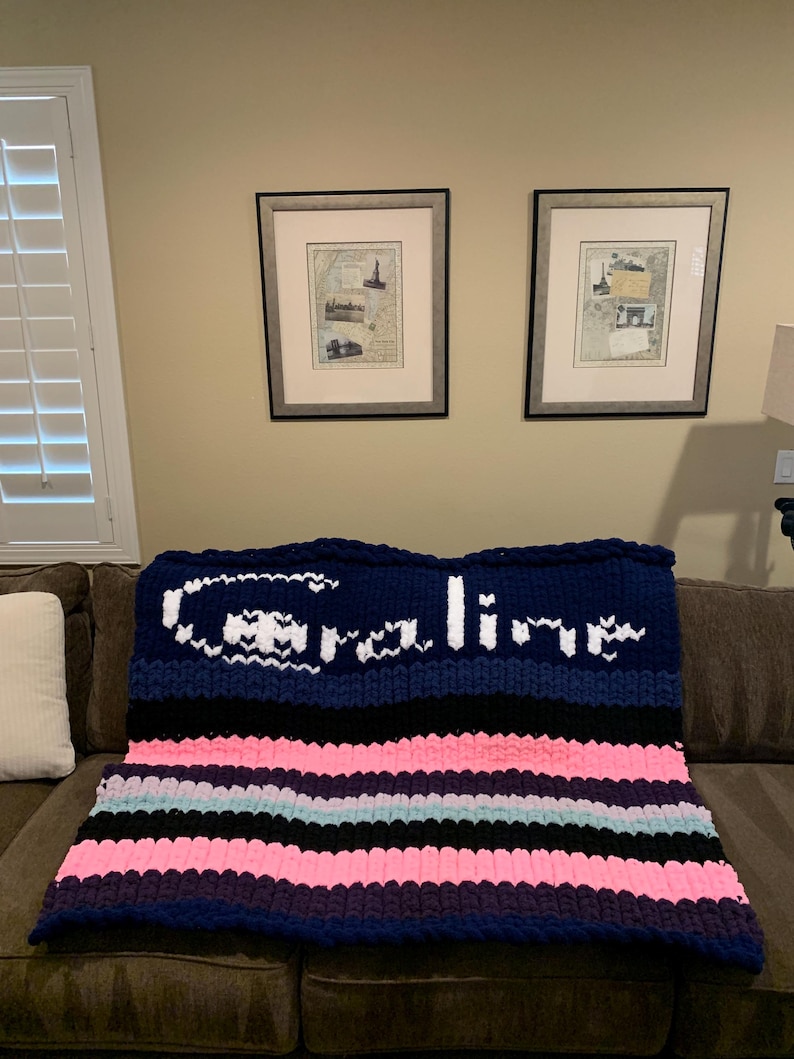K&ouml;nnte beinhalten: Eine grob gestrickte Decke mit marineblauen, schwarzen, rosa und hellblauen Streifen. Der obere Bereich zeigt den Namen "Caroline" in wei&szlig;en Buchstaben. Die Decke liegt auf einem braunen Sofa und schafft eine gem&uuml;tliche Atmosph&auml;re.