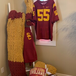 May include: A display of collegiate fan gear in cardinal red and gold. Items include a football jersey with the number 55, a chunky knit blanket, a sweatshirt, a t-shirt with "Southern California Trojans" text, and a football.