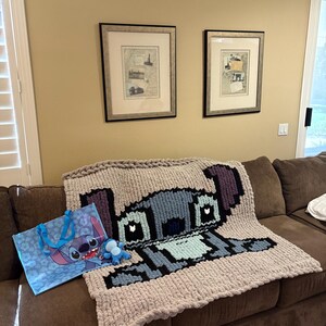 Puede incluir: Una manta grande de punto grueso con un dise&ntilde;o pixelado del personaje Stitch de Lilo & Stitch. La manta es principalmente gris claro con acentos azules, negros y morados. Tambi&eacute;n se ve una bolsa de mano a juego y un peque&ntilde;o peluche.