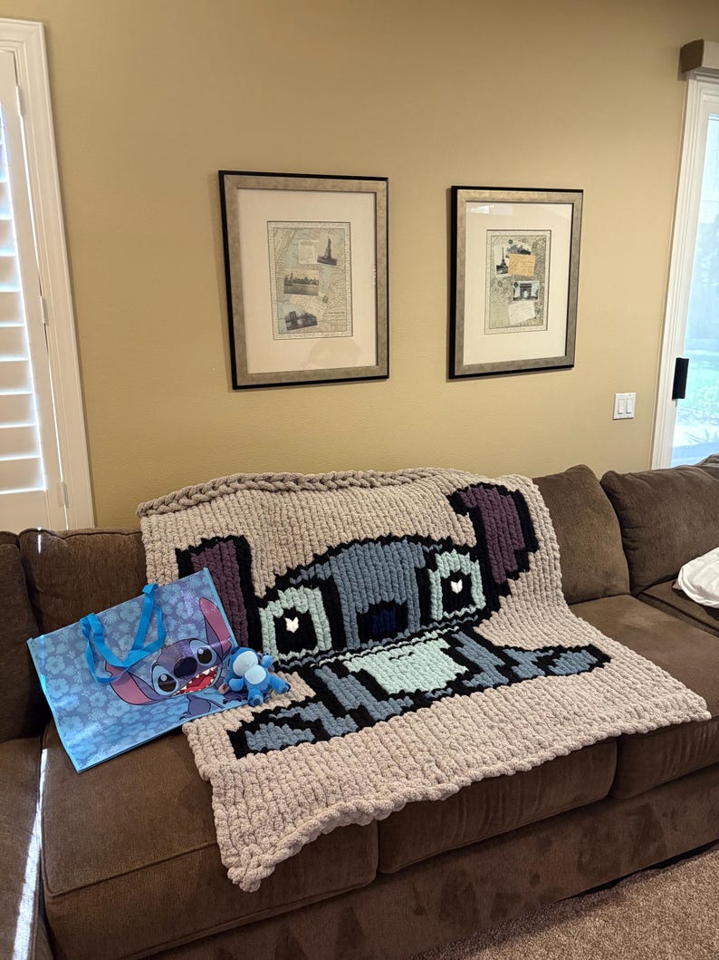 Puede incluir: Una manta grande de punto grueso con un dise&ntilde;o pixelado del personaje Stitch de Lilo & Stitch. La manta es principalmente gris claro con acentos azules, negros y morados. Tambi&eacute;n se ve una bolsa de mano a juego y un peque&ntilde;o peluche.