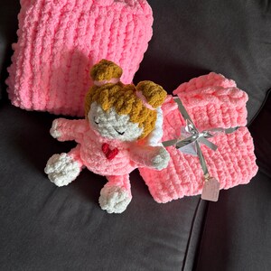 Peut inclure: Un ensemble coussin et couverture rose en tricot &eacute;pais avec une poup&eacute;e crochet&eacute;e. La poup&eacute;e a un visage blanc, des cheveux bruns et un c&oelig;ur rouge. La couverture est attach&eacute;e avec un ruban argent&eacute;. Les articles sont sur un canap&eacute; gris fonc&eacute;.