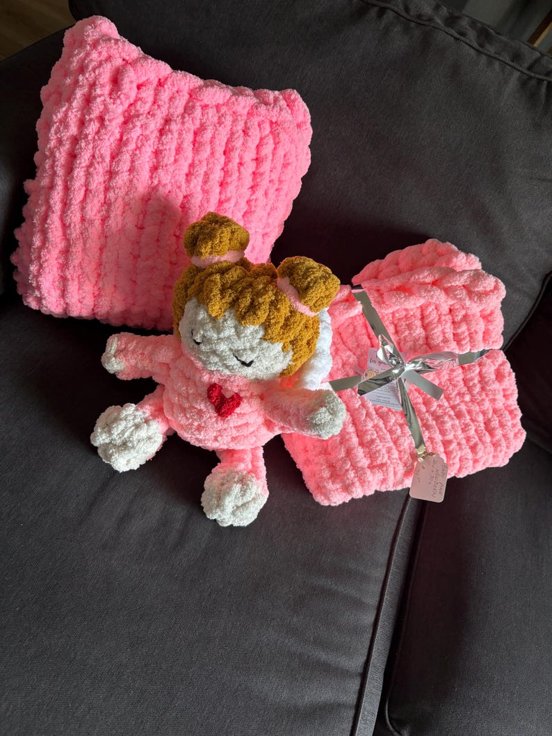 Peut inclure: Un ensemble coussin et couverture rose en tricot &eacute;pais avec une poup&eacute;e crochet&eacute;e. La poup&eacute;e a un visage blanc, des cheveux bruns et un c&oelig;ur rouge. La couverture est attach&eacute;e avec un ruban argent&eacute;. Les articles sont sur un canap&eacute; gris fonc&eacute;.
