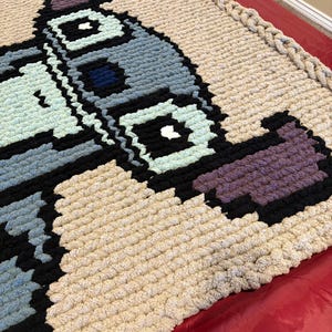 Puede incluir: Una manta pixelada grande, hecha a mano, en una variedad de colores. La manta presenta un dise&ntilde;o con tonos de azul, negro, morado y verde claro sobre un fondo beige. Los bordes est&aacute;n acabados con un borde grueso y trenzado.