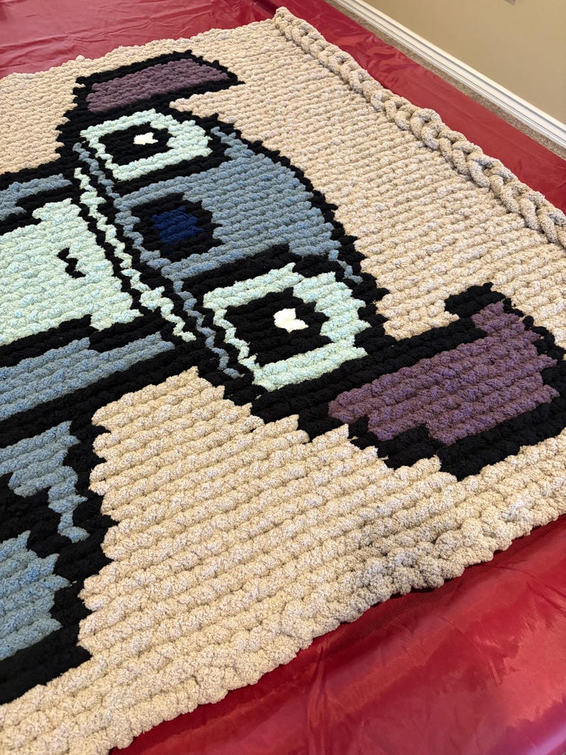 Puede incluir: Una manta pixelada grande, hecha a mano, en una variedad de colores. La manta presenta un dise&ntilde;o con tonos de azul, negro, morado y verde claro sobre un fondo beige. Los bordes est&aacute;n acabados con un borde grueso y trenzado.