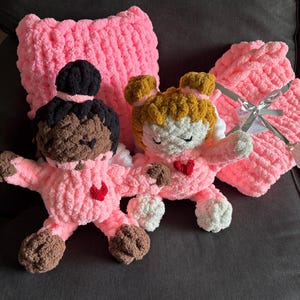 Peut inclure: Deux poup&eacute;es roses faites &agrave; la main au crochet, avec des cheveux bruns et noirs, portant des tenues roses avec des c&oelig;urs rouges. Un oreiller et une couverture roses au crochet sont derri&egrave;re les poup&eacute;es. Les poup&eacute;es ont des ailes blanches.