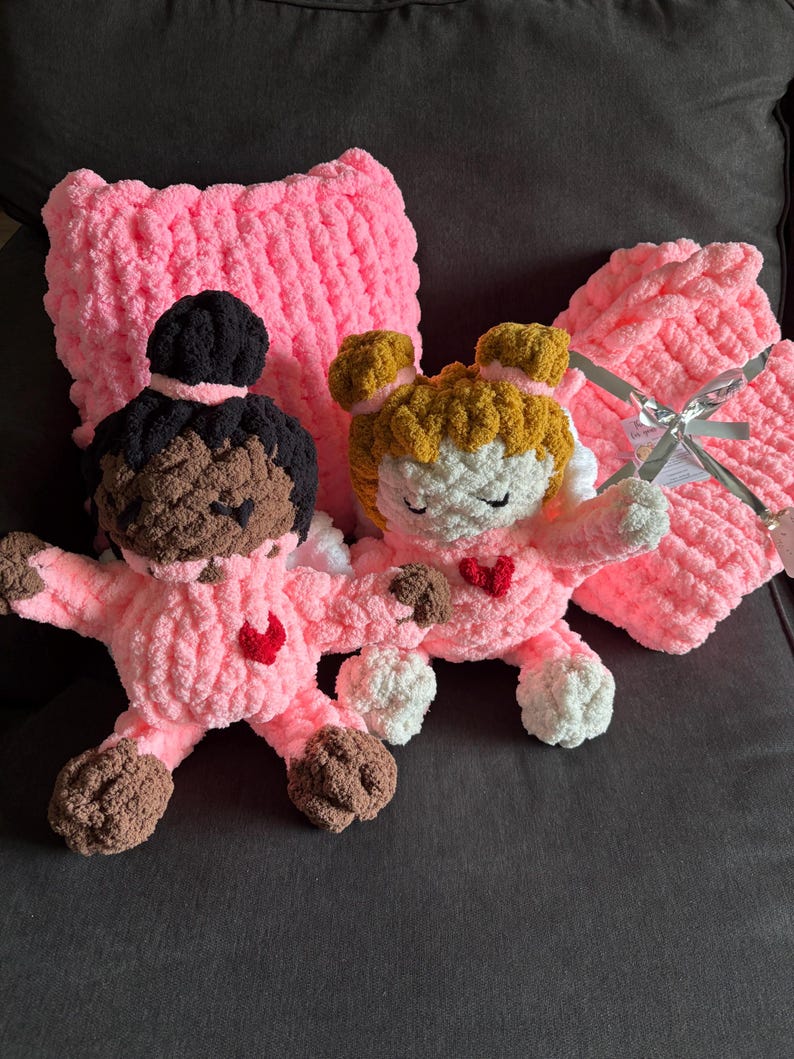 Peut inclure: Deux poup&eacute;es roses faites &agrave; la main au crochet, avec des cheveux bruns et noirs, portant des tenues roses avec des c&oelig;urs rouges. Un oreiller et une couverture roses au crochet sont derri&egrave;re les poup&eacute;es. Les poup&eacute;es ont des ailes blanches.