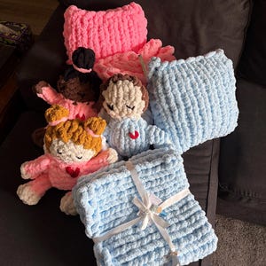 Peut inclure: Ensemble d'articles faits &agrave; la main au crochet, comprenant des coussins et des couvertures roses et bleus. Plusieurs petites poup&eacute;es au crochet avec diff&eacute;rentes couleurs de peau et de cheveux sont &eacute;galement pr&eacute;sentes, chacune avec un c&oelig;ur rouge.