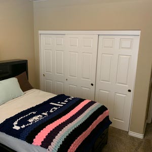 K&ouml;nnte beinhalten: Eine grob gestrickte Decke mit dem Namen "Caroline" in wei&szlig;en Buchstaben auf marineblauem Hintergrund. Die Decke hat Streifen in Rosa, Schwarz und Hellblau. Die Decke liegt auf einem Bett mit schwarzem Rahmen, in einem Schlafzimmer mit wei&szlig;en Schrankt&uuml;ren.