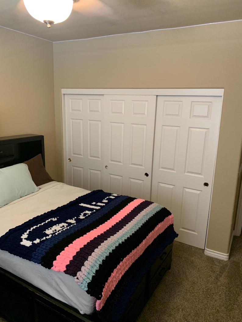 K&ouml;nnte beinhalten: Eine grob gestrickte Decke mit dem Namen "Caroline" in wei&szlig;en Buchstaben auf marineblauem Hintergrund. Die Decke hat Streifen in Rosa, Schwarz und Hellblau. Die Decke liegt auf einem Bett mit schwarzem Rahmen, in einem Schlafzimmer mit wei&szlig;en Schrankt&uuml;ren.