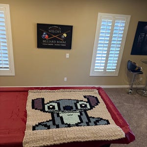 Puede incluir: Una manta de felpa hecha a mano con una imagen pixelada del personaje Stitch de Lilo & Stitch. La manta es beige con detalles en azul, gris y negro, y se muestra sobre una mesa de billar roja.