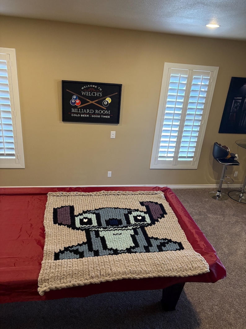 Puede incluir: Una manta de felpa hecha a mano con una imagen pixelada del personaje Stitch de Lilo & Stitch. La manta es beige con detalles en azul, gris y negro, y se muestra sobre una mesa de billar roja.